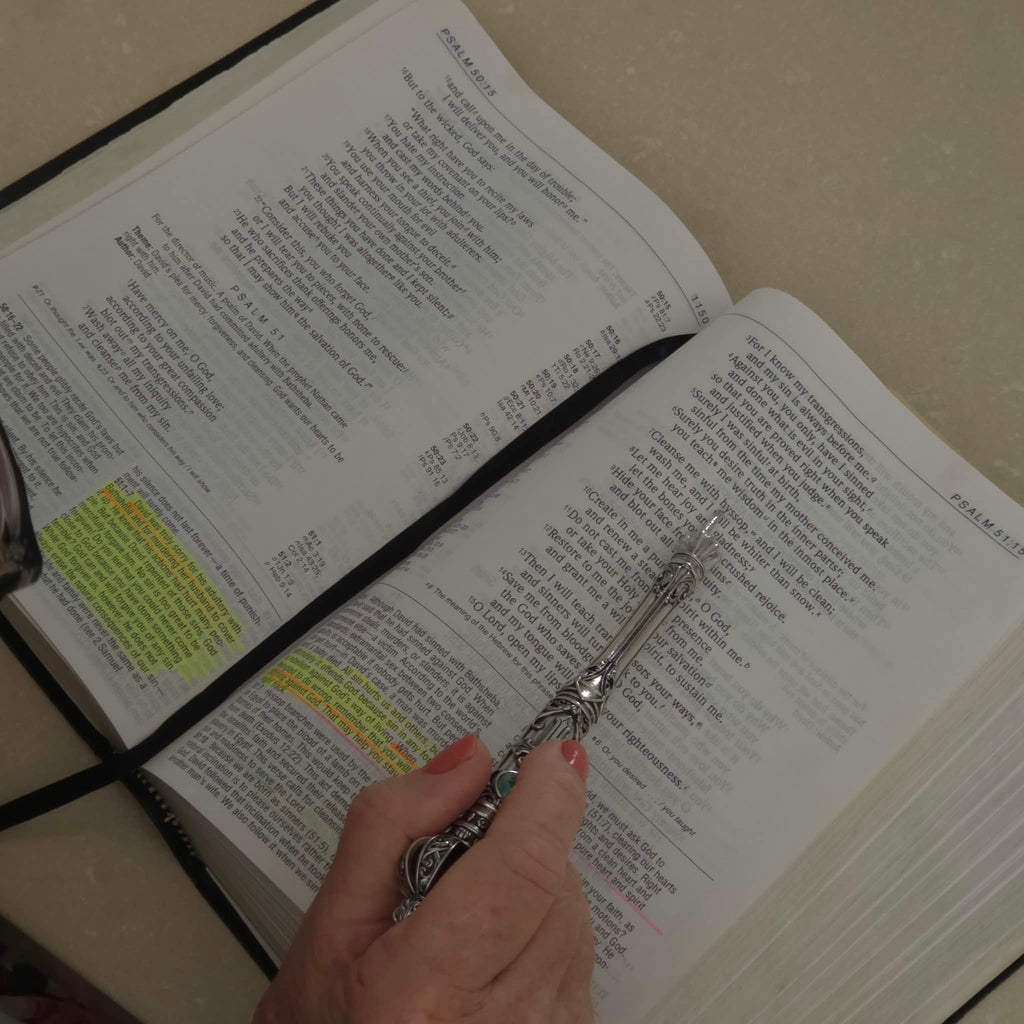 A hand holding the Moriah Cross Works Bible Reading Pointer in bright silver, pointing to scripture text open at Psalm 51, with highlighted verses visible on the page