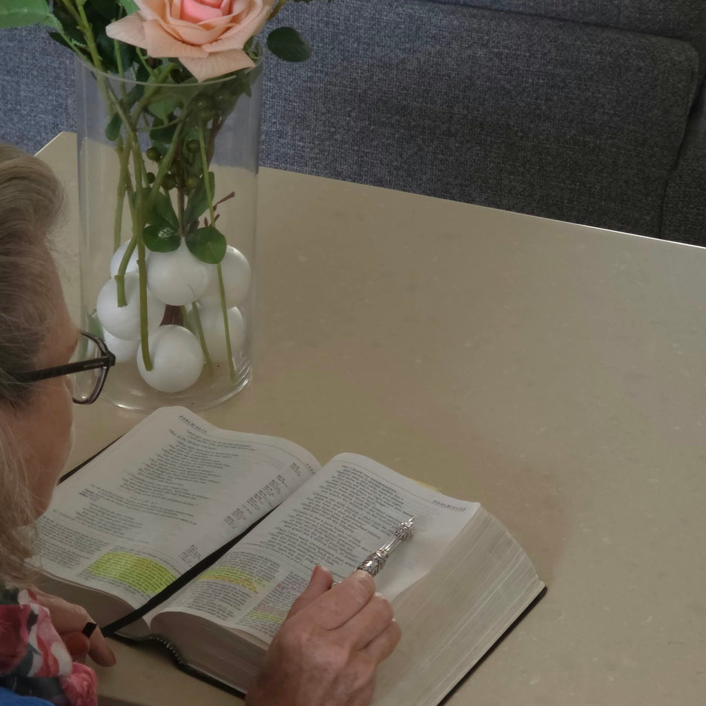 An elderly woman reading an open Bible at a table, using the Moriah Cross Works Bible Reading Pointer in bright silver to follow the scripture text, photographed from above with a vase of roses visible beside the Bible