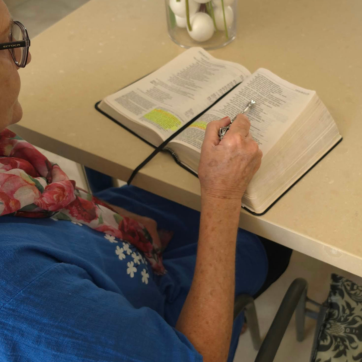 An elderly woman seated at a table reading an open Bible, using the Moriah Cross Works Bible Reading Pointer to follow the text, photographed from behind and above showing the full reading context